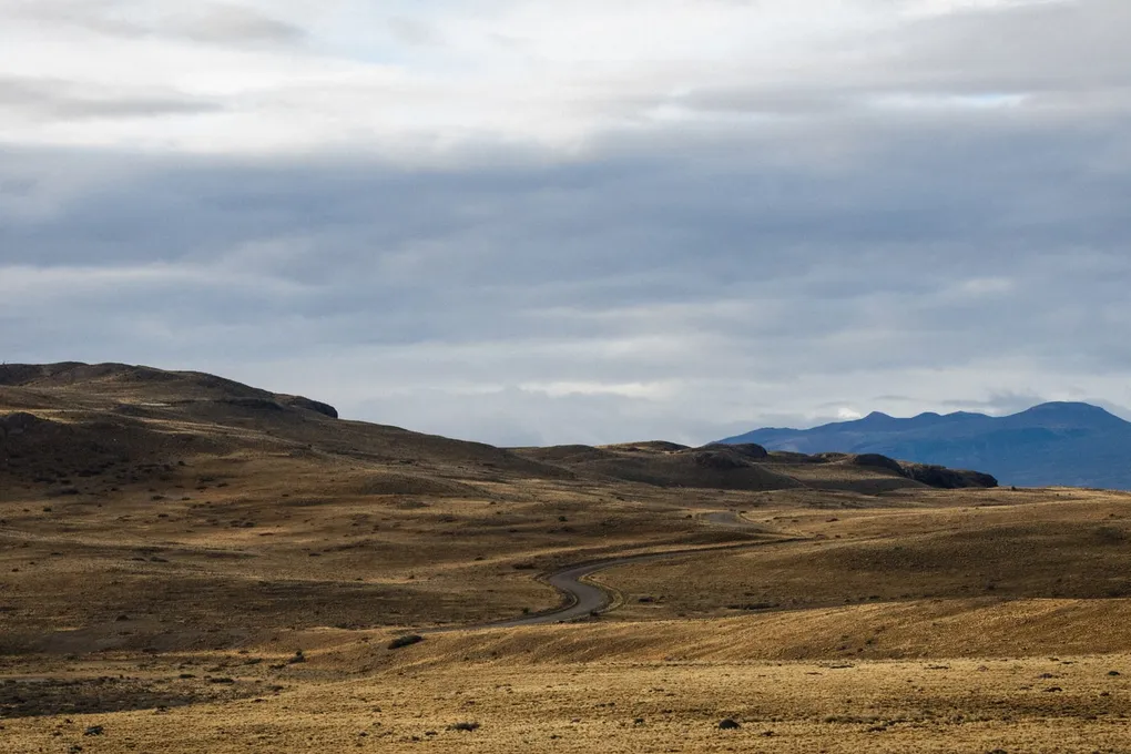 A wide, arid landscape of rolling brown hills with a winding road cutting through the terrain, set against distant mountains under a cloudy sky.