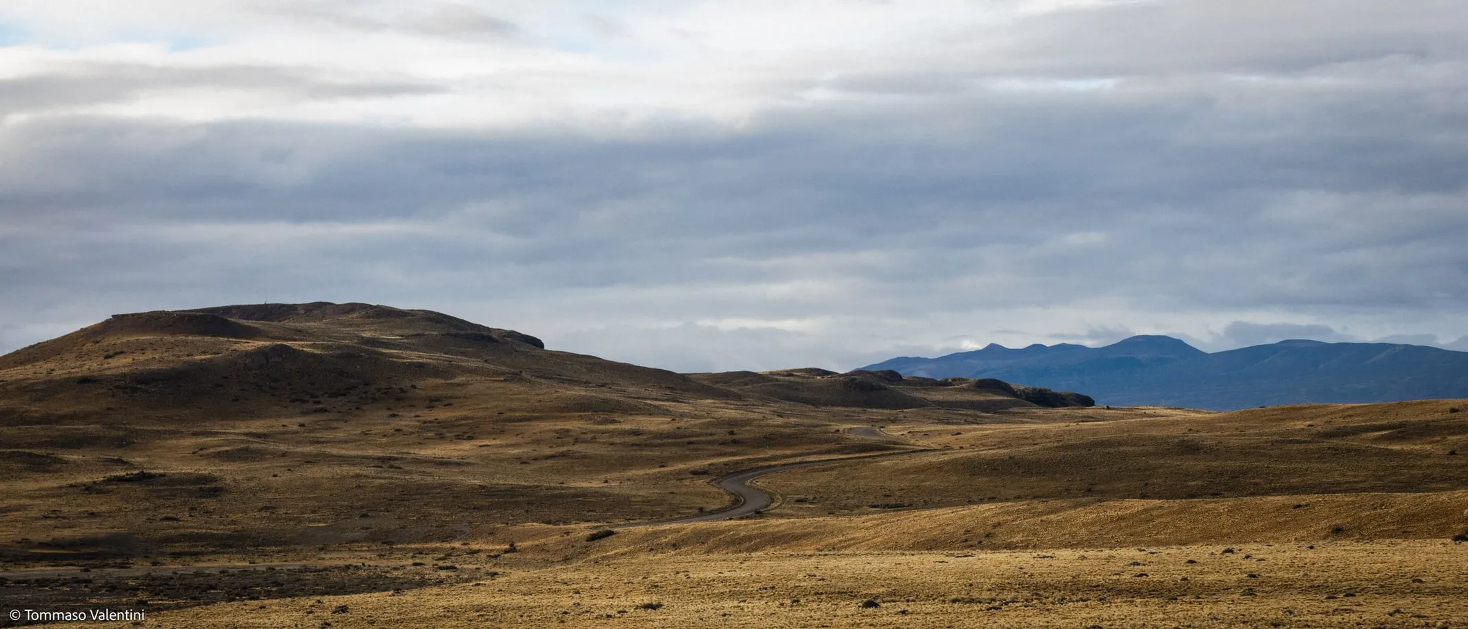 A wide, arid landscape of rolling brown hills with a winding road cutting through the terrain, set against distant mountains under a cloudy sky.
